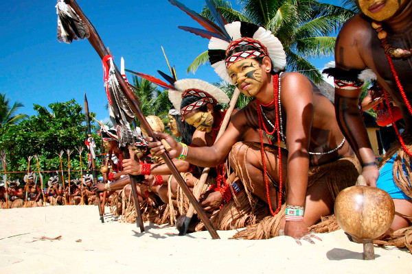 Indígenas agachados com suas vestimentas típicas e rosto pintado, uma cultura celebrada no Dia dos Povos Indígenas.