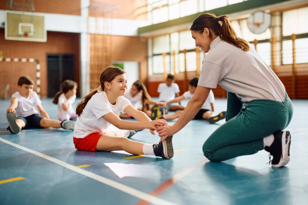 Professora dando aula de alongamento, durante a disciplina de Educação Física, para uma turma de crianças em um ginásio.